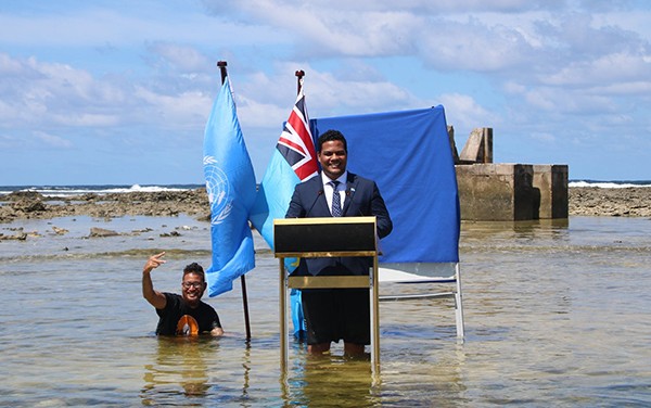 Kofe stands at a lectern in the shallow water, flanked by UN flags.