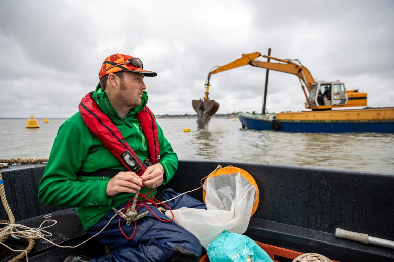 Tom Cameron sitting in a boat off the coast of Essex, UK, watching a digger on a nearby barge drop stone and shell into the sea