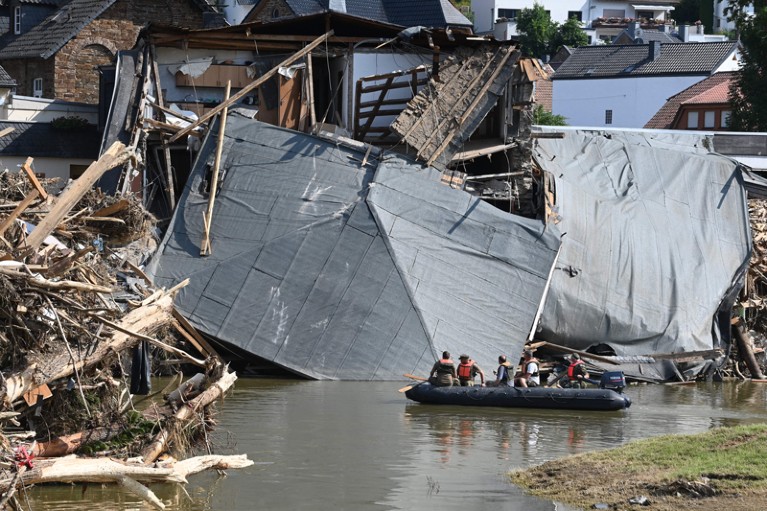 Military personnel floats on a boat on a river as the roof of a damaged house hangs in the water