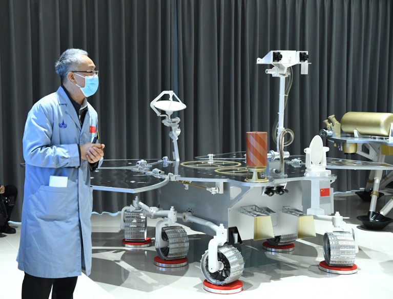 A man in a mask and lab coat next to a model rover.