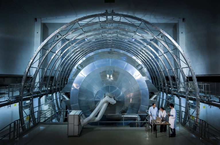 Three people in lab coats stand next to the metal chamber in which electrons are measured as part of the KATRIN experiment.