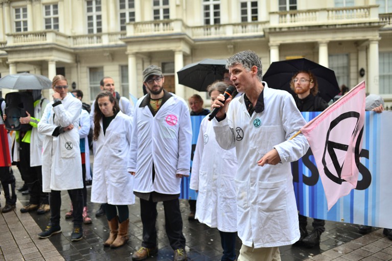 A line of people in lab coats standing outside in the rain. One man speaks into a microphone.