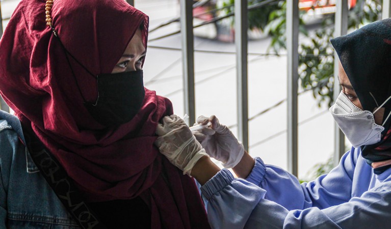 A woman in protective clothing and a head scarf injects another woman in a head scarf and amsk.