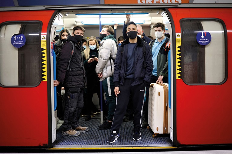 Crowded tube train with door open, showing commuters squeezed together, wearing masks.
