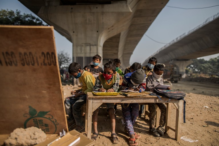 在一个建筑工地的临时教室里，孩子们戴着口罩坐在立交桥下的一张桌子前