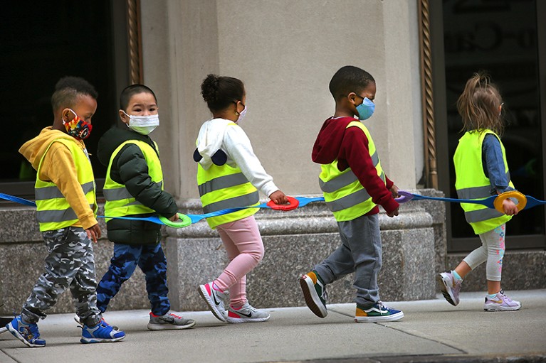 Children in a preschool class wear vests and masks and hold on to a strap while walking on a street in Boston.