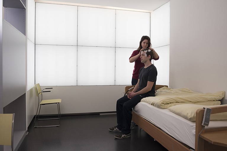 An employee attaches electrodes to a man’s head as he sits on a bed. Behind them panelled lights can be seen on the wall.