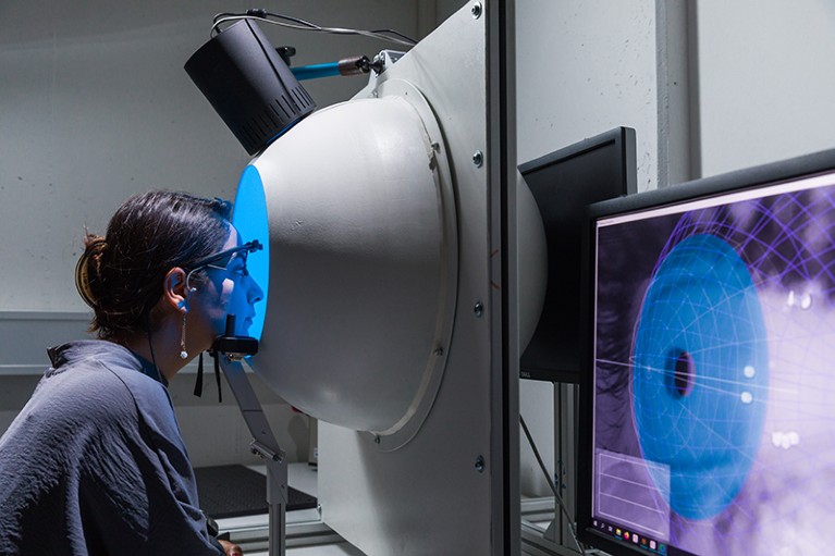A woman wearing an eye apparatus stares into a machine. In the foreground a computer shows a closeup of her eye.