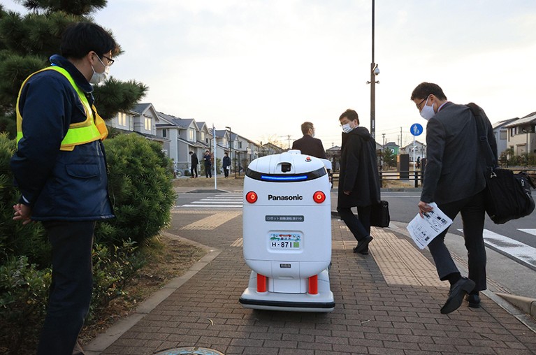 Three people wearing face masks look at a delivery robot on a street at the Fujisawa Sustainable Smart Town