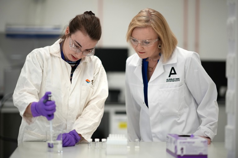 Liz Truss wears a white lab coat and protective glasses while watching a scientist carry out an experiment