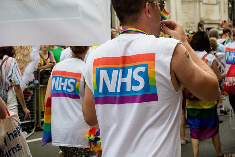 在骄傲游行中，一名NHS工作人员穿着一件背心，上面的NHS标志叠加在骄傲旗上
