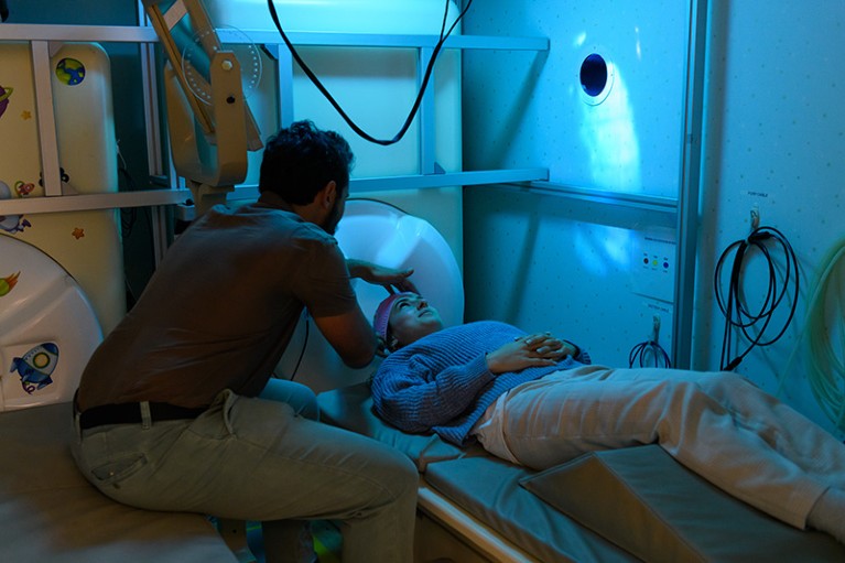 Vince Polito adjusts the cap of a woman lying down in a blue lit room - behind her is a large plastic MEG scanner