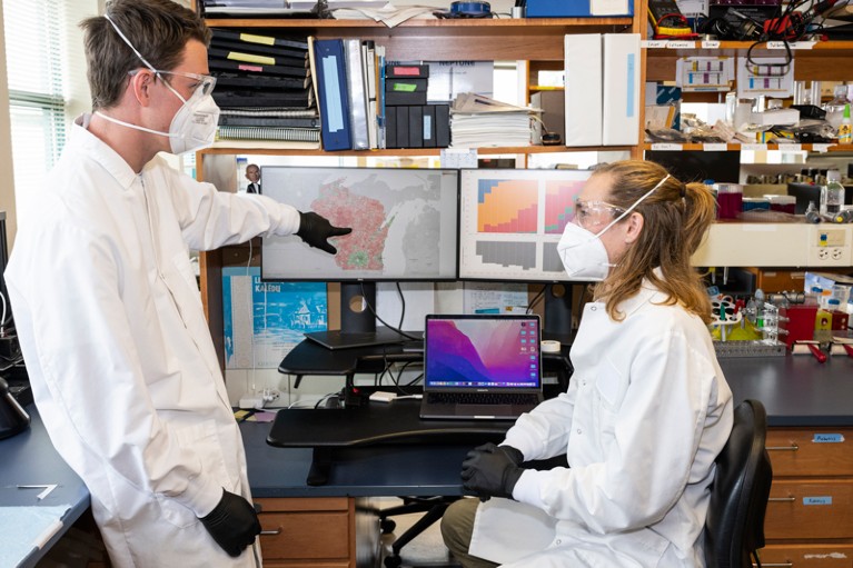 Two scientists wearing masks, goggles and lab coats point at a computer screen displaying data