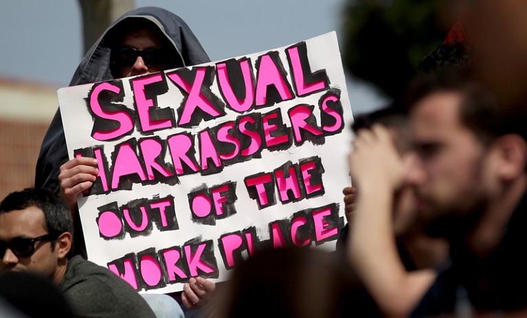 A demonstrator wearin a hood and sunglasses holds a sign reading "Sexual harrassers out of the workplace" in bright pink letters