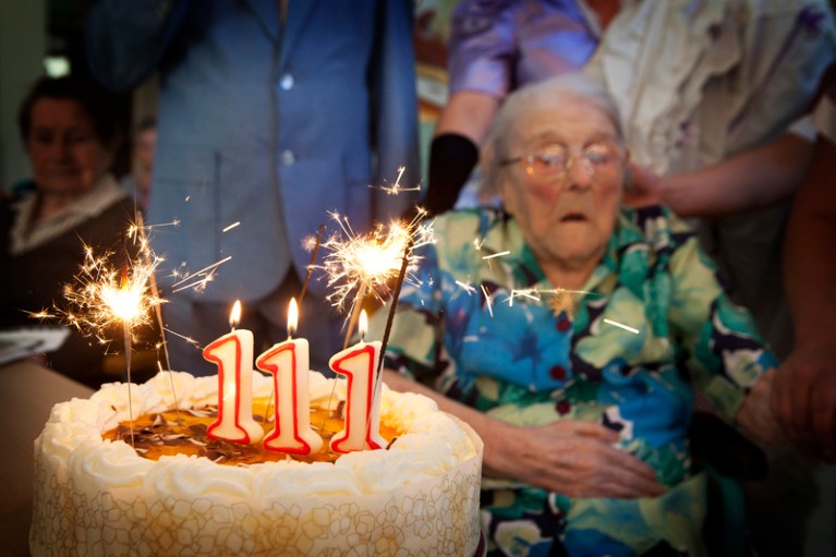 An elderly woman, surrounded by people, sitting behind a birthday cake with sparklers and candles spelling 111.