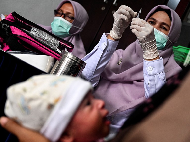 A woman in a headscarf holds a syringe. Another person holds a baby in the foreground, and a third stands behind.