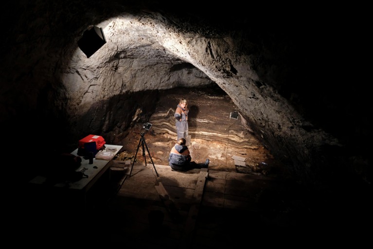 A light illuminates part of a stratified wall and two archaeologists working in the Denisova Cave in Siberia.