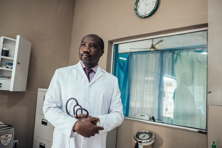Dimie Ogoina wearing a white coat and holding a stethoscope posing for a portrait in a doctors surgery