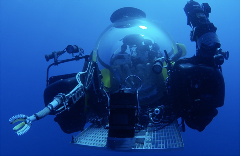 A Triton 3300 submarine is pictured under water with a soft robotic arm to the left of the photo