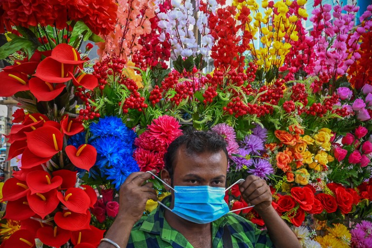A vendor selling artificial flowers puts on his face mask with a wall of colourful flowers behind him.