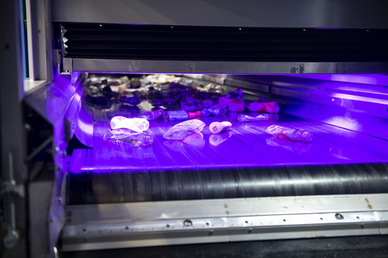 A machine with a conveyor belt has various recycling packages on it, bathed in an ultraviolet light.
