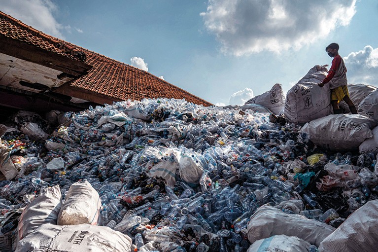 A young boy wearing a mask stands on a huge pile of plastic waste. He is holding a large white sack.