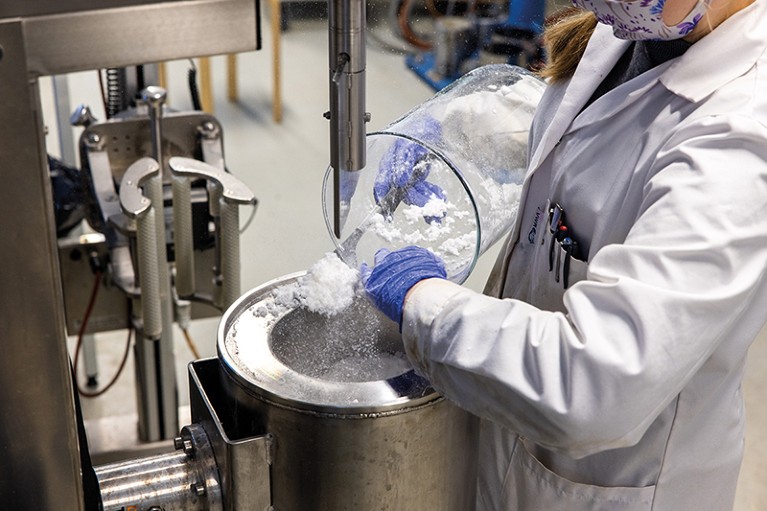 A woman in white lab coat and mask pouring white, fluffy substance from a container into a metal receptacle.