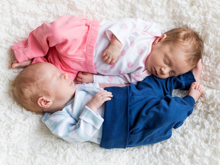 Newborn twins sleeping on white blanket, one dressed in blue and one in pink.