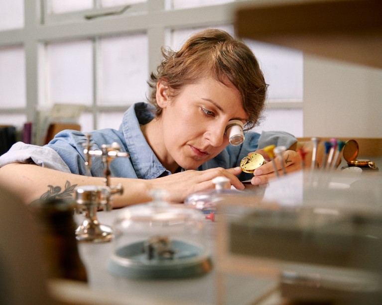 Rebecca Struthers at a workbench covered in tools in her workshop inspects a golden watch with a magnifying eyeglass