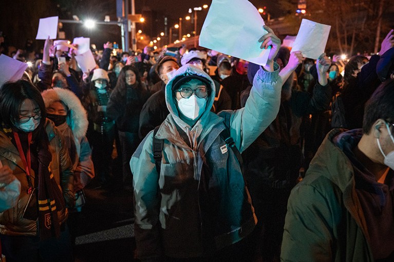 A crowd of people, most wearing face masks and many holding blank pieces of paper.