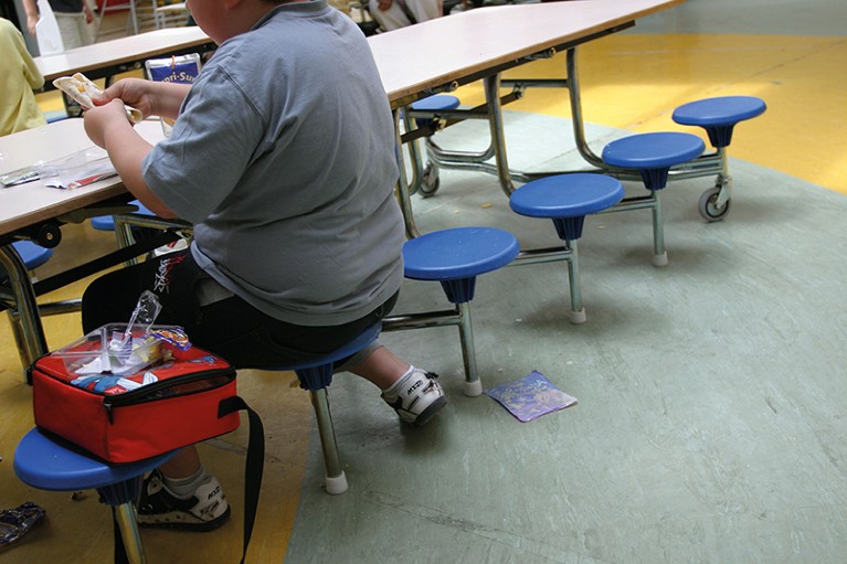Mid section view of a boy sitting at a school dining table. He is overweight and is eating a wrap.