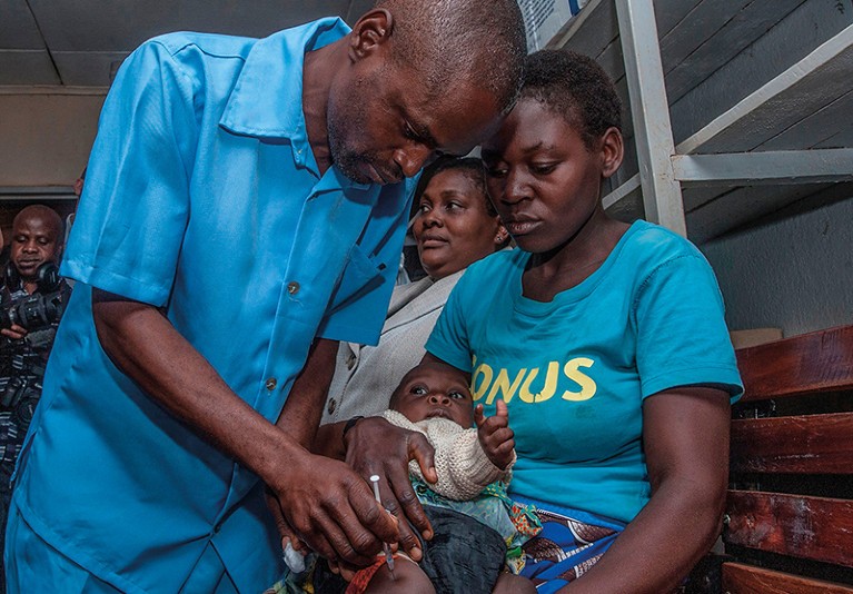 A man wearing a blue shirt injects a vaccine into the thigh of a baby. The baby is held by its mother.