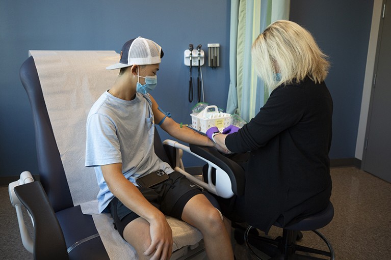 Rear view of a seated blonde woman taking blood from the arm of a young man.