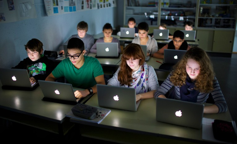 Rows of pupils work on laptops at desks in a dark classroom.