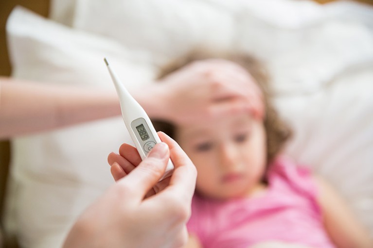 Sick child with high fever laying in bed and mother holding thermometer with temperature reading of 38.9 degrees celsius