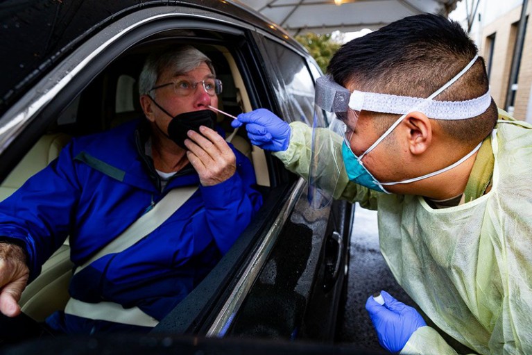 A healthcare professional wearing PPE administers a COVID-19 test to a patient in his car