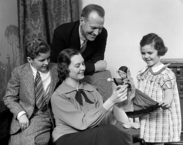 A black and white photo of a mother, father, son and daughter playing with a toy doll.