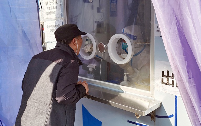 A person in protective gear inside a kiosk swabs the mouth of a customer.
