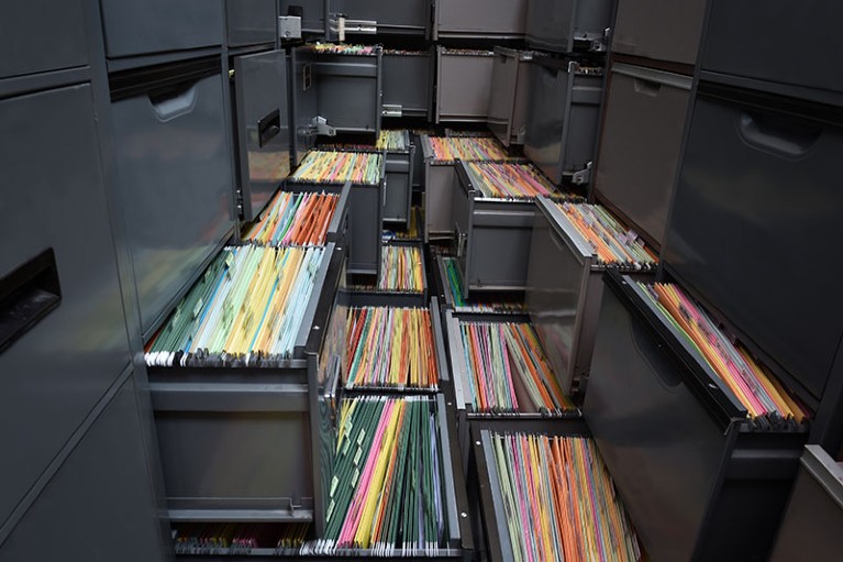 A tightly packed cluster of filing cabinets with some drawers open exposing their contents of documents.