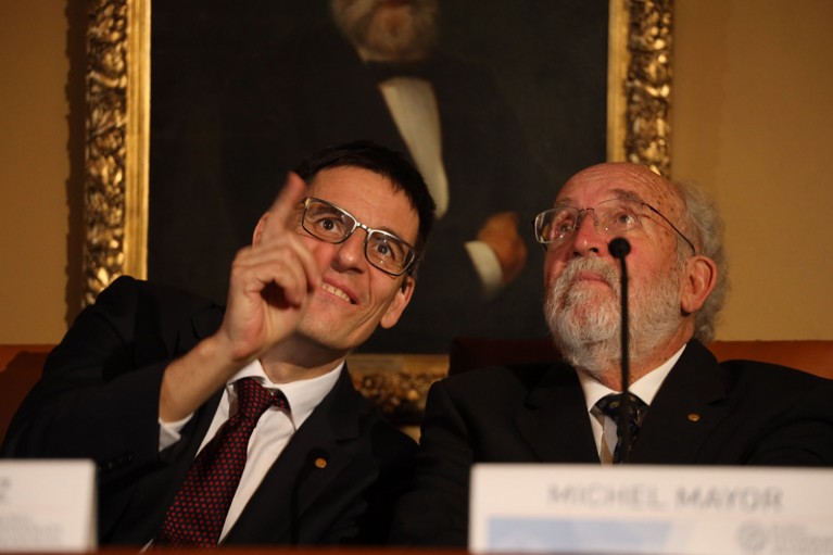 Didier Queloz and Michel Mayor sitting together and looking at something out of frame.