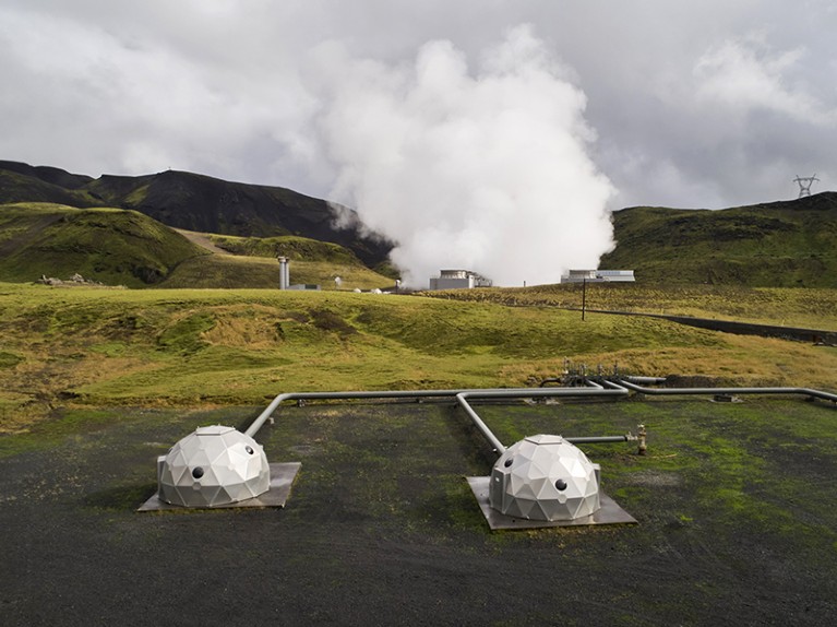 Silvery pods connected to pipes, in front of green hills from which a cloud is billowing.