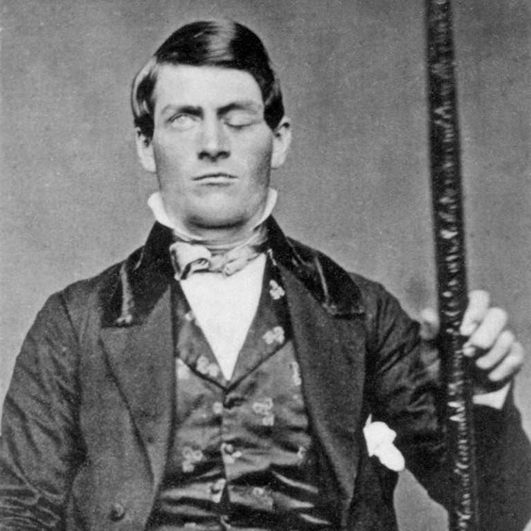 Black and white photo of a man in nineteenth-century clothing, with one eye closed, holding a metal rod.