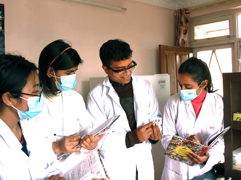 Four people in white coats stand in a lab. One of them is holding a small device, and the others are writing in notebooks.