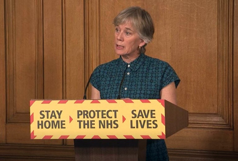 Angela McLean stands behind a podium labelled 'Stay home, protect the NHS, save lives'.