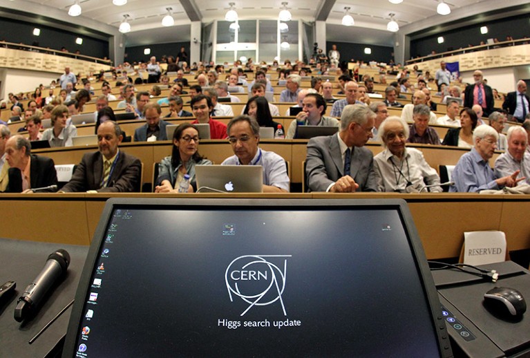 View from a lectern looking into a full lecture theatre, a computer monitor displays the words Higgs search update.