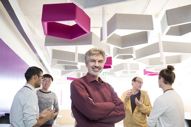Yoshua Bengio satnds in front of a group of four adults who are discussing things in pairs.