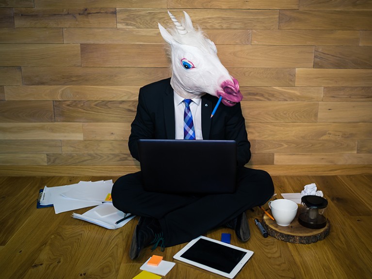Young man in funny horse mask sits on the floor near a wall and works with laptop.