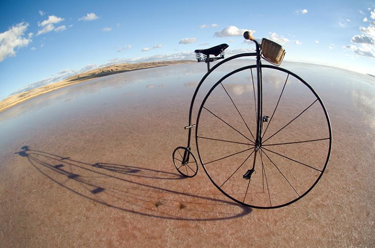 A penny farthing antique bicycle on wet sand in front of blue sky.