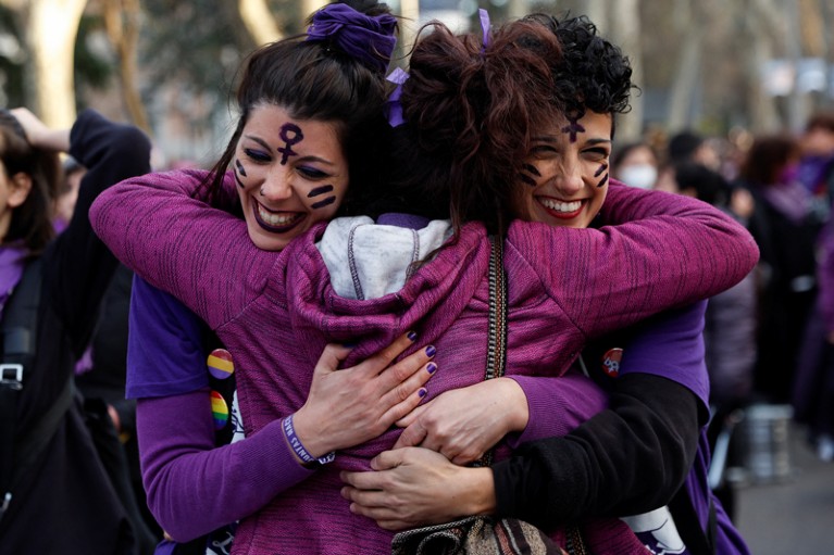 Three women smiling and wearing purple clothes embrace at an International Women's Day protest