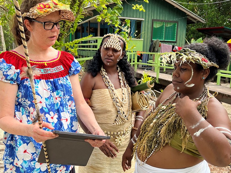 One woman in a blue dress holds an iPad, showing it to two women wearing tan clothes, headpieces and many necklaces.